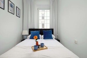 Ein ordentlich bezogenes Bett mit blauen Kissen, ein Tablett mit Tassen und einer Blume, und ein Fenster mit Vorhängen.