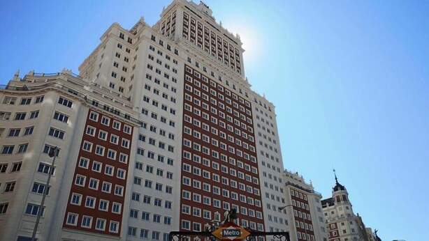 Un hotel de varios pisos con una fachada destacada de color rojo y blanco, un cartel de Metro que indica acceso al transporte público y una torre del reloj.