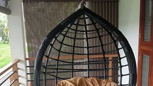 Una silla colgante con un cojín marrón en una habitación con suelo de baldosas, baranda de madera y ventana con persianas.
