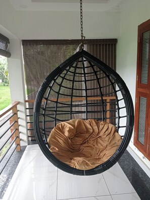 Una silla colgante con un cojín marrón en una habitación con suelo de baldosas, baranda de madera y ventana con persianas.