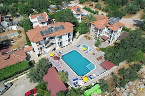 Une vue aérienne d’un quartier résidentiel avec une piscine entourée de chaises longues et de parasols, des panneaux solaires sur les toits et un bâtiment au toit rouge.