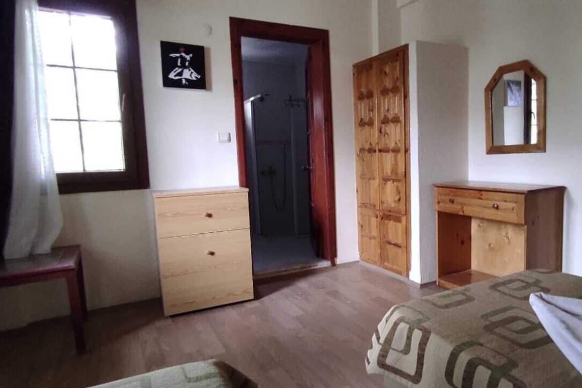 A bedroom with a wooden wardrobe, a wooden dresser, a mirror, a bed with a blanket, and a window with curtains.