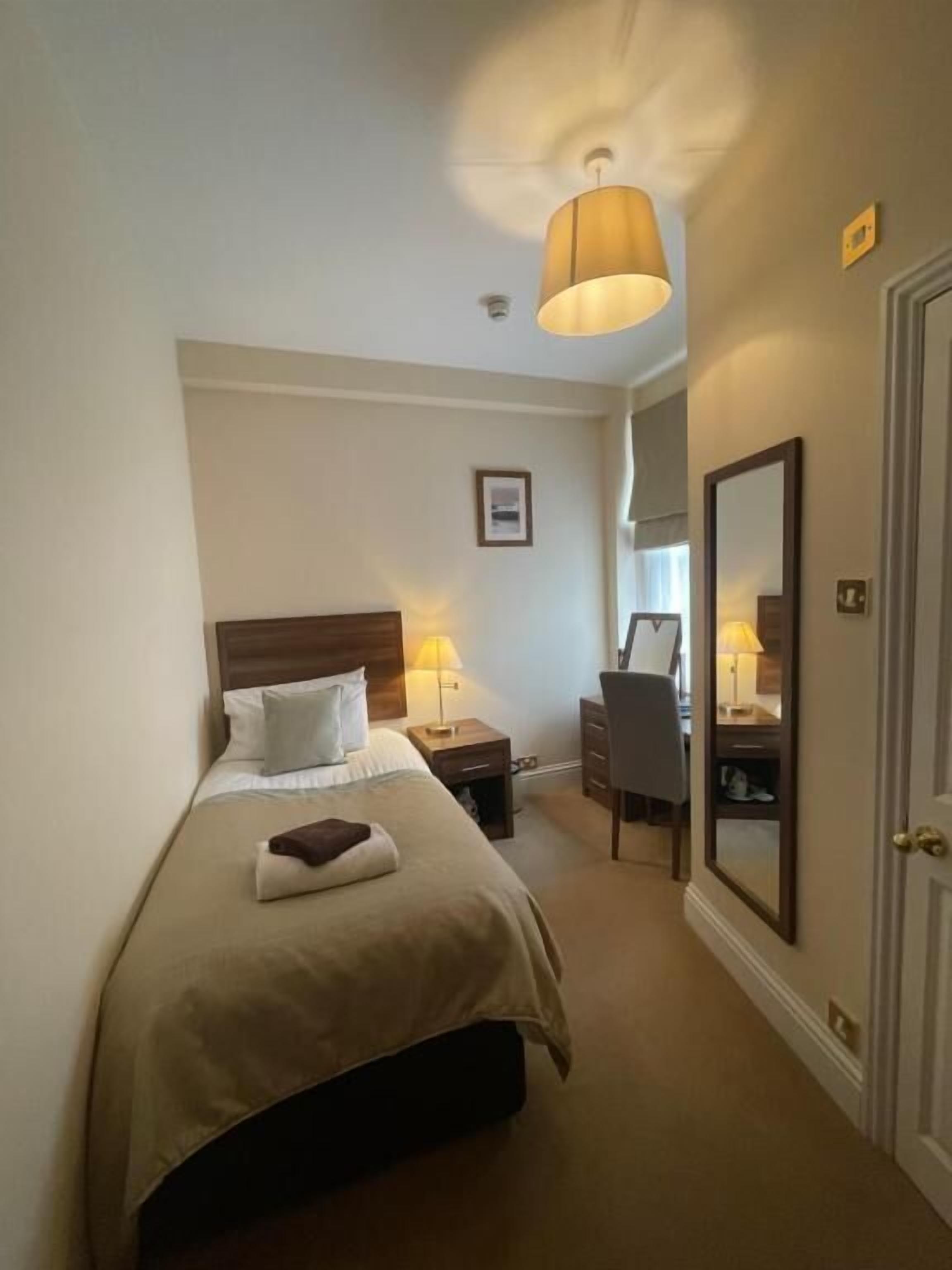 A hotel room with a bed, bedside table, lamp, desk, chair, mirror, and a door.