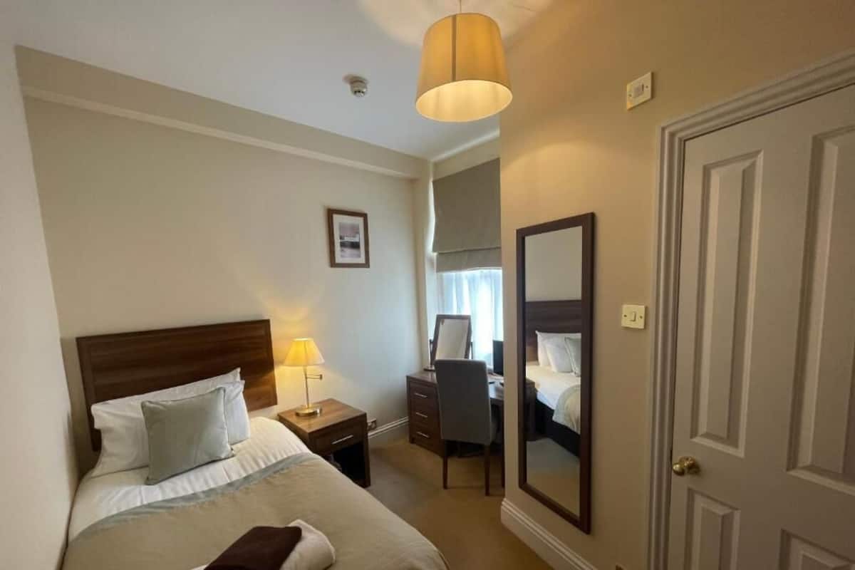A hotel room with a bed, bedside table, lamp, desk, chair, mirror, and a door.