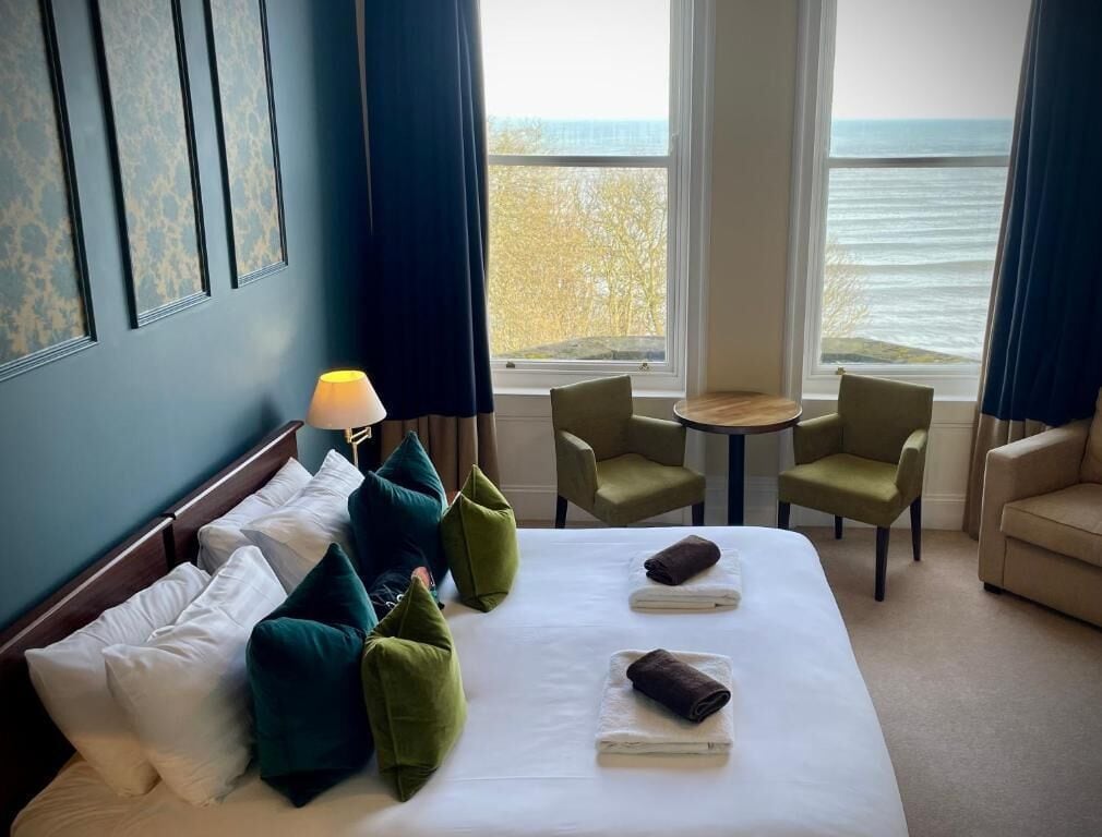 Habitació d'hotel amb un llit, cadires verdes, una taula petita i vista a l'oceà.