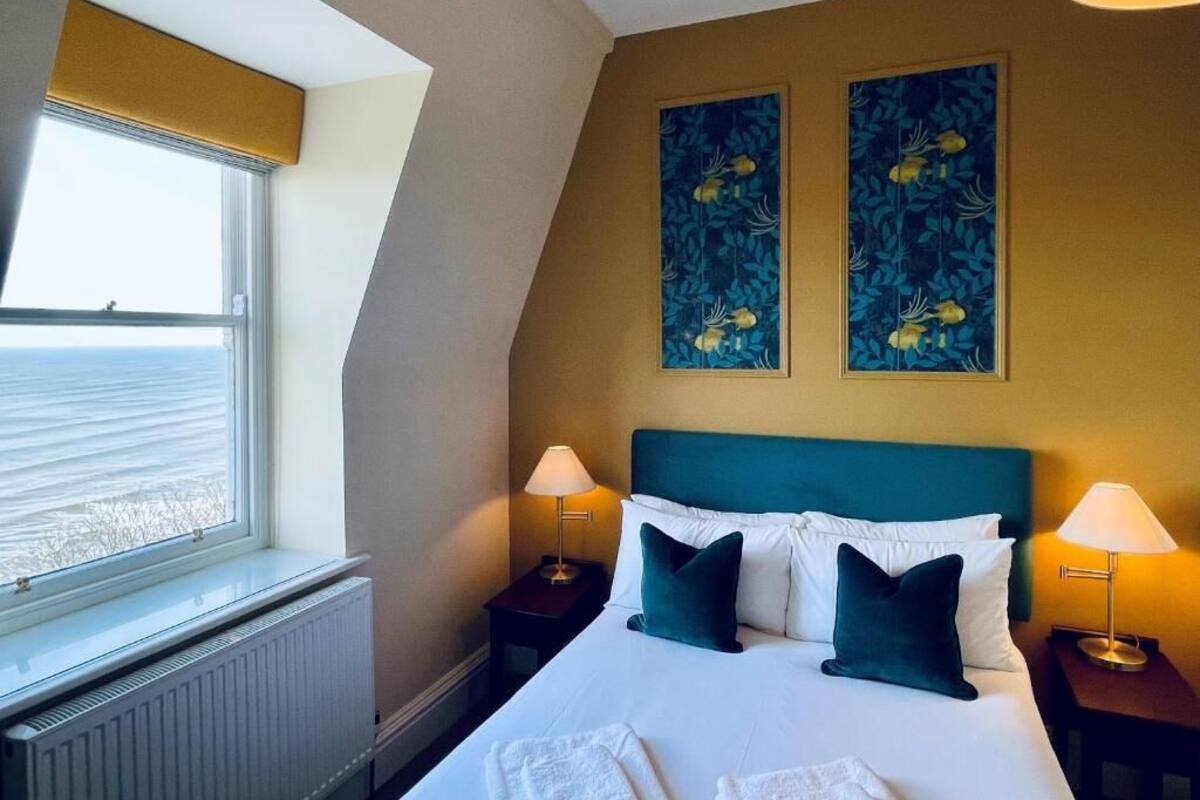A hotel room with a bed, two bedside lamps, a window with a view, and two wall art pieces.