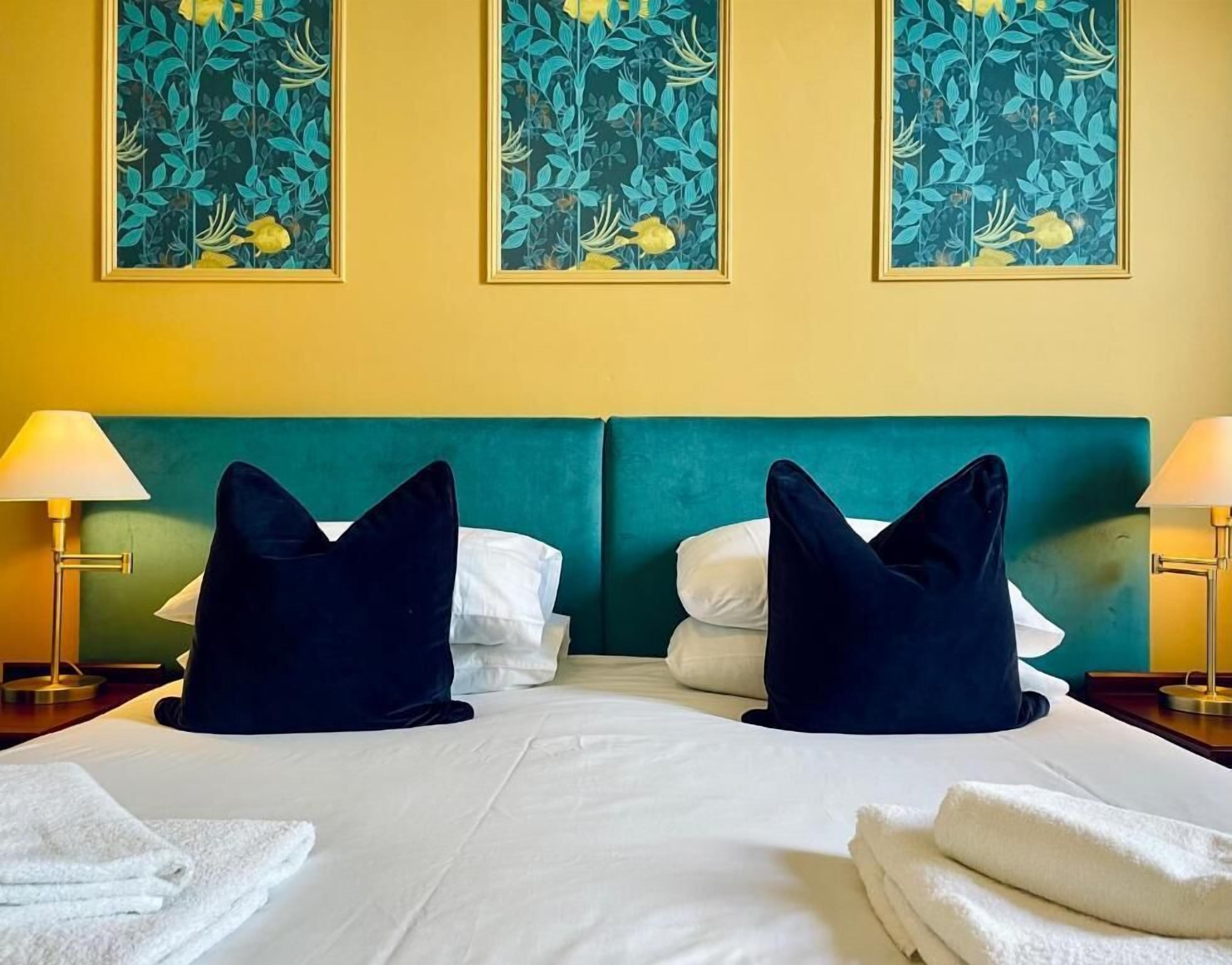 Habitación de hotel con una cama, dos almohadas, un cabecero, dos lámparas de noche y una pared decorada con tres cuadros de flores.