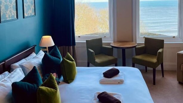 Habitación de hotel con cama, almohadas, lámpara, mesa, sillas y ventanas amplias con vista al mar.