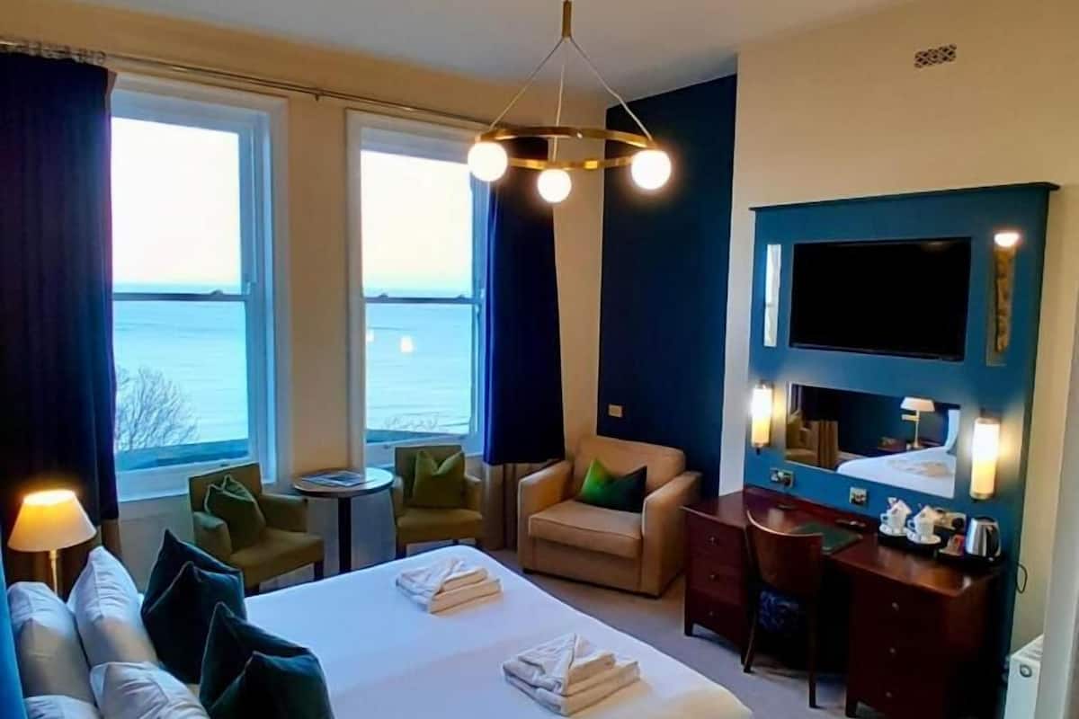 A hotel room with a bed, a desk, a chair, a TV, and a view of the sea.
