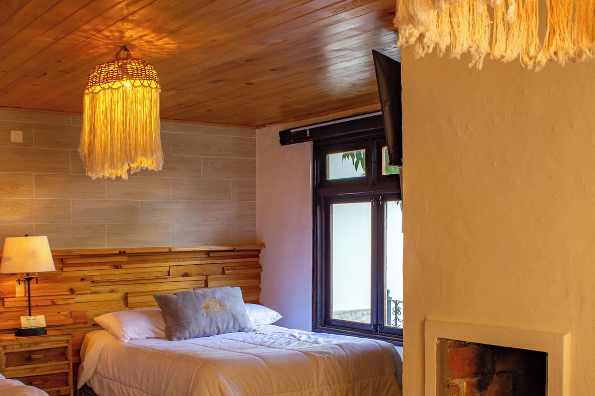 A bedroom with a wooden ceiling, a bed with a headboard, a nightstand with a lamp, a fireplace, and a window with a view.