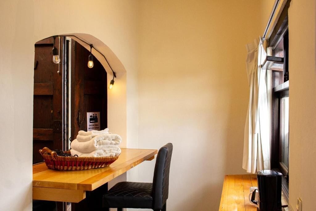 Une petite pièce avec une table en bois, une chaise noire, un panier rempli de serviettes, une fenêtre avec des rideaux et un panneau accroché au mur.