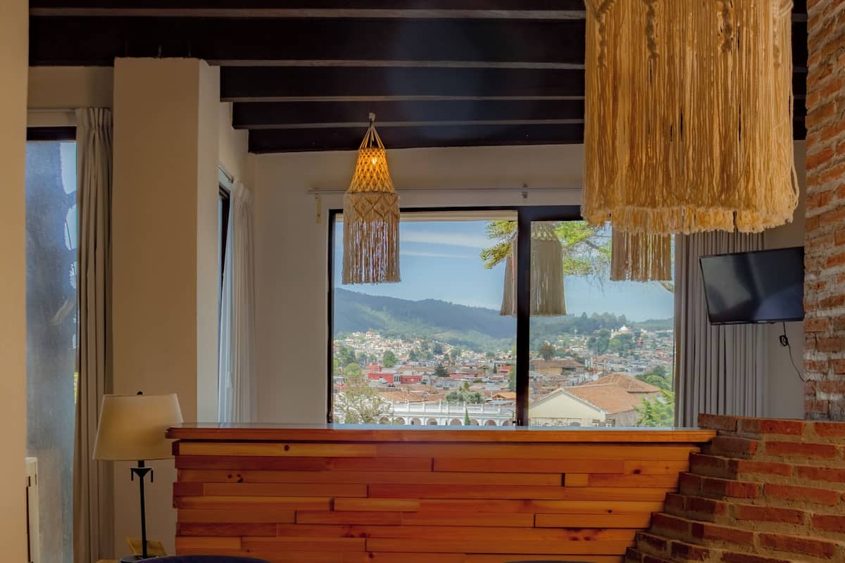 A room with a wooden bench, two chairs, a hanging light fixture, a TV, and a view of a town through a window.