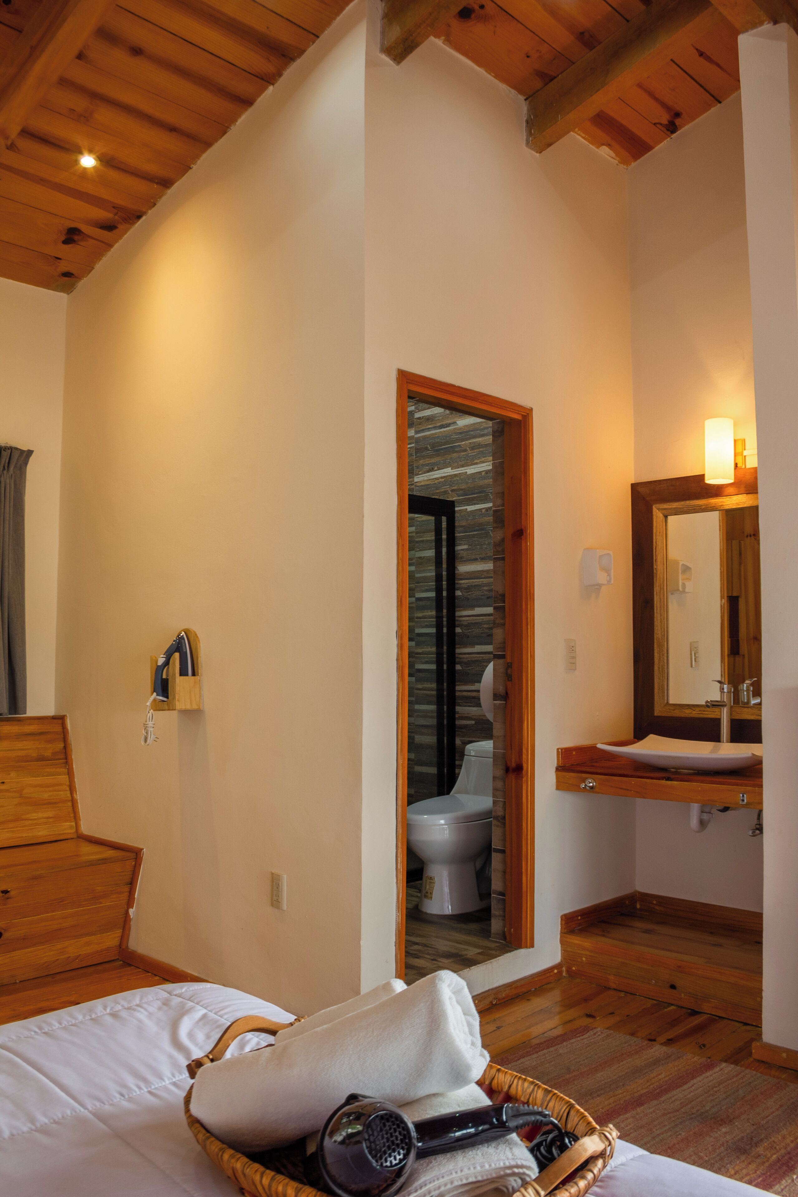 A room with a wooden ceiling, a bed with a white comforter, a wicker basket with towels and a hairdryer, a wooden dresser, and a bathroom with a wooden vanity and mirror.
