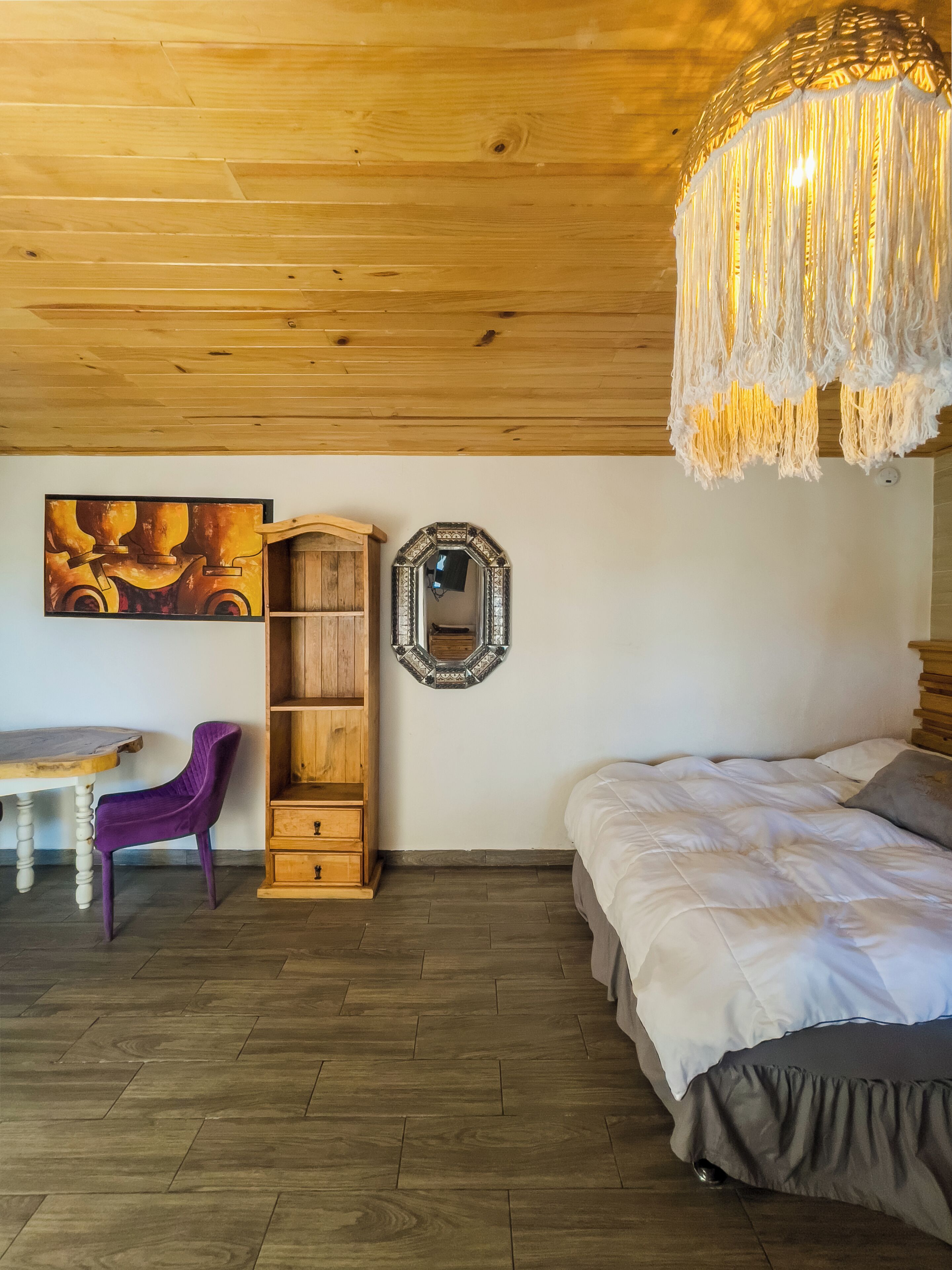 Un dormitorio con techo de madera, una cama, un armario de madera, un espejo, una mesa y una silla morada.
