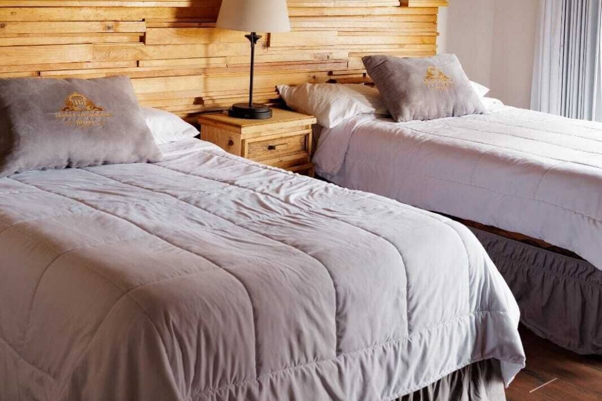 A hotel room with two beds, a wooden headboard, a nightstand with a lamp, and a window with curtains.