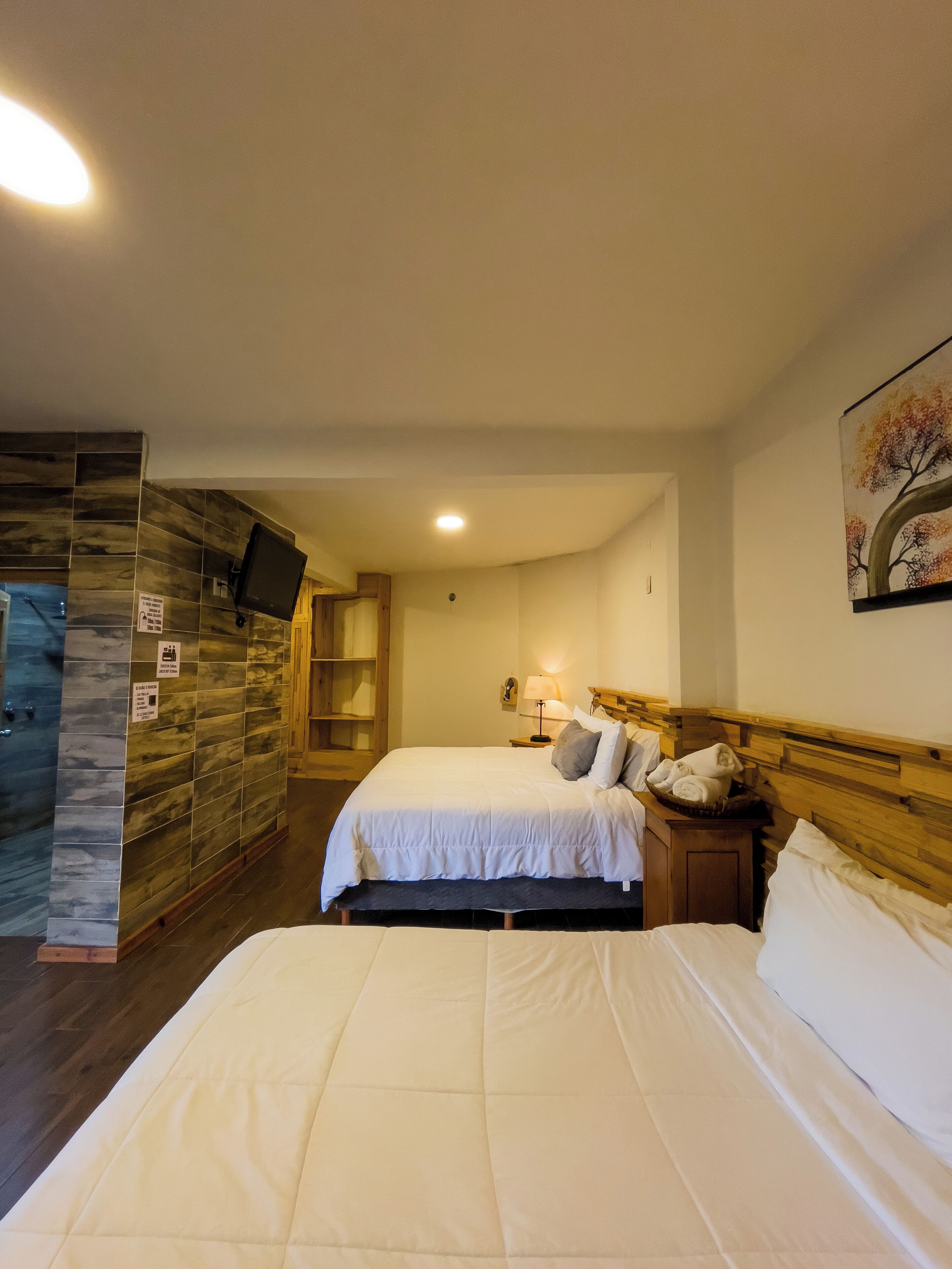 Ein Hotelzimmer mit zwei Betten, einem an der Wand befestigten Fernseher, einem Gemälde und einem hölzernen Kopfteil.