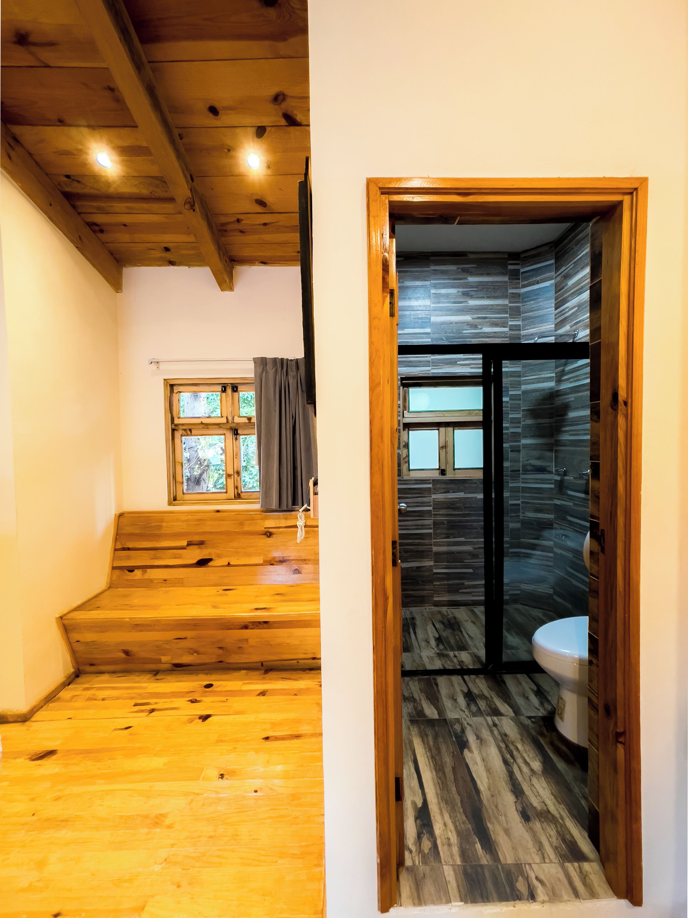 Un escalier en bois mène à une pièce dotée d’une fenêtre, d’un espace douche avec une porte en verre et de toilettes.