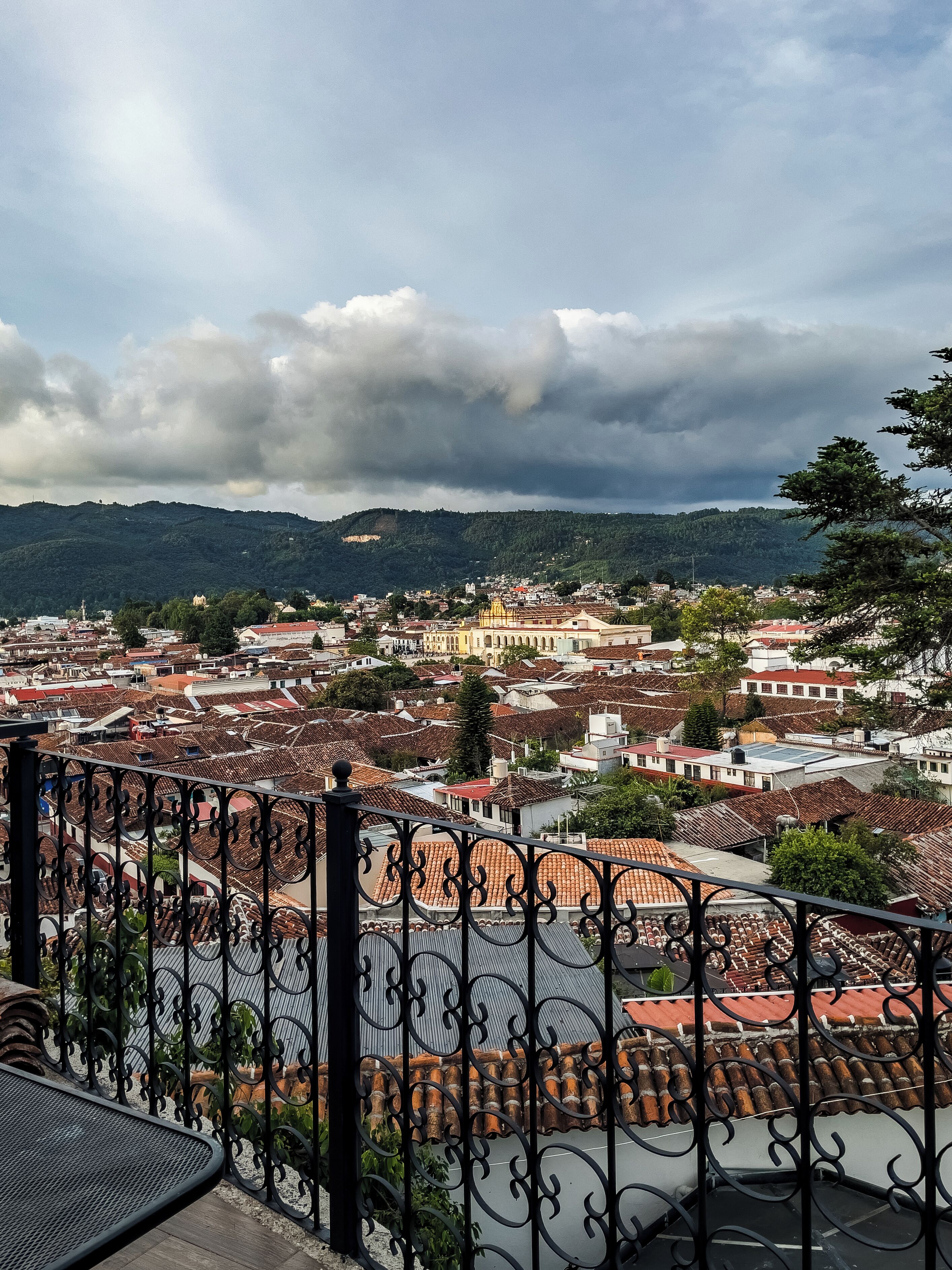 A cityscape with red-tiled rooftops, a balcony with ornate black railings, and a cloudy sky.