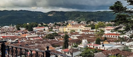 A cityscape with red-tiled rooftops, a balcony with ornate black railings, and a cloudy sky.