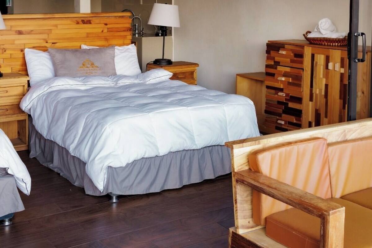 A bedroom with a wooden headboard, a bed with white bedding, a hanging macrame light fixture, a wooden nightstand, a bedside table with a lamp, and a wooden dresser.