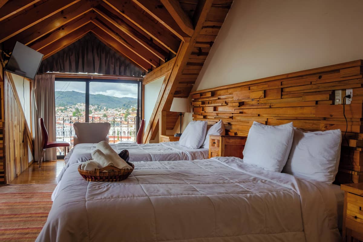 A room with two beds, a wooden headboard, a window with a view of a town, and a red chair.
