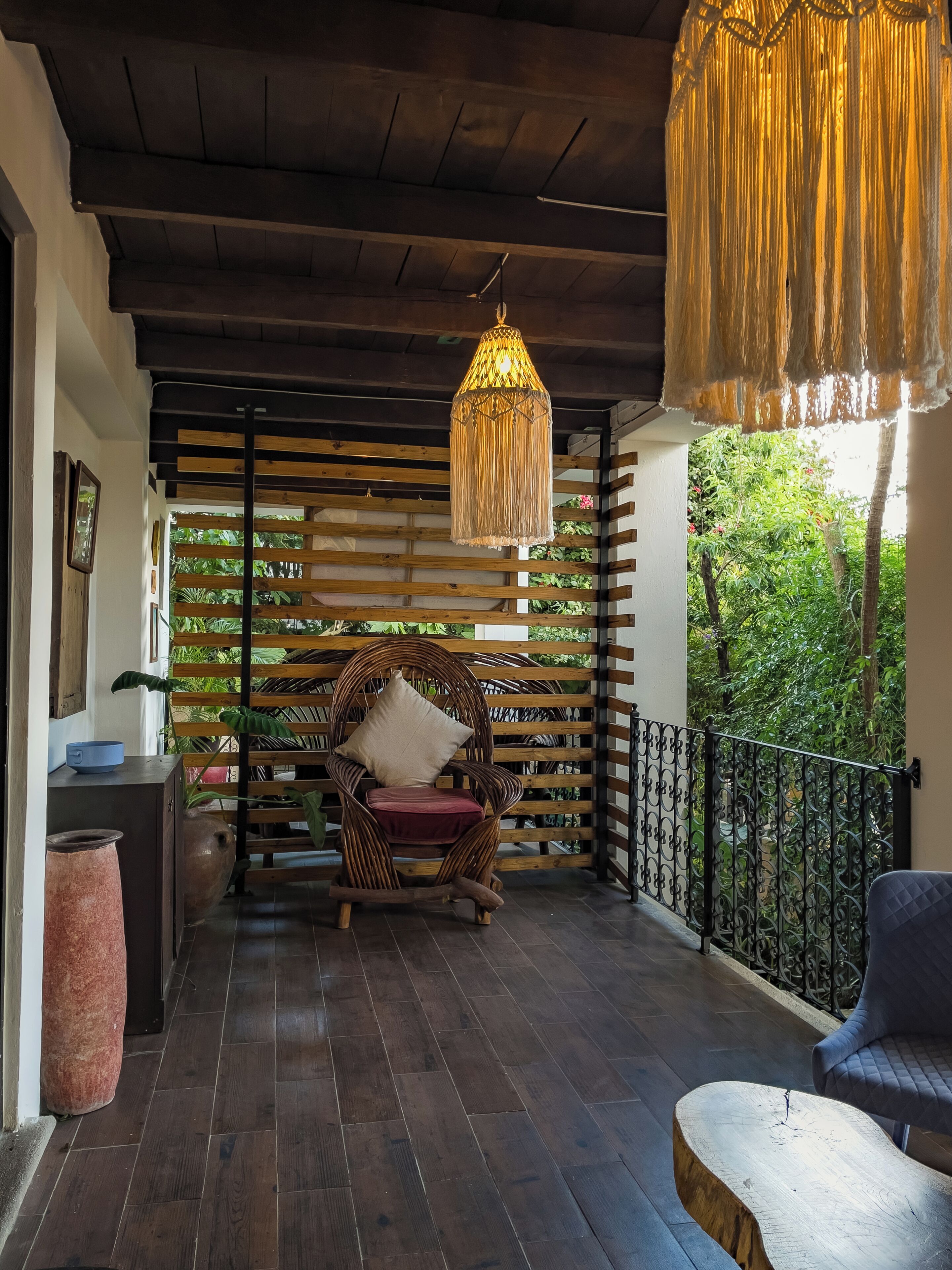A wooden ceiling, a wicker chair with a cushion, a hanging light fixture, and a view of greenery.