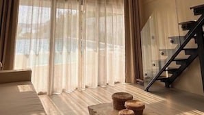 Una habitación con un ventanal grande, cortinas traslúcidas, un ventilador de techo, una escalera y una mesa de madera con taburetes.