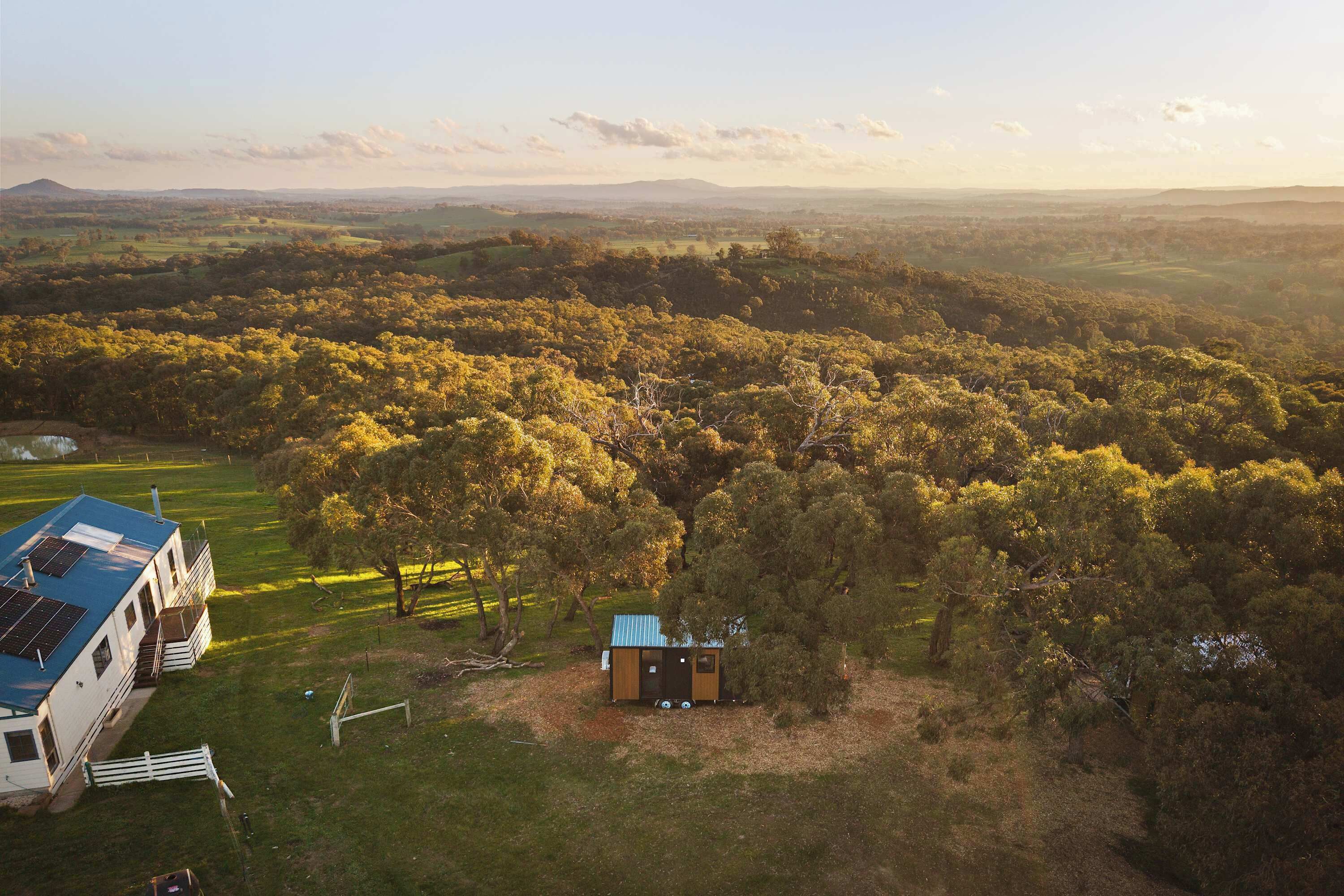 A rural landscape with a house featuring a blue roof and solar panels, surrounded by dense forest and rolling hills in the background.