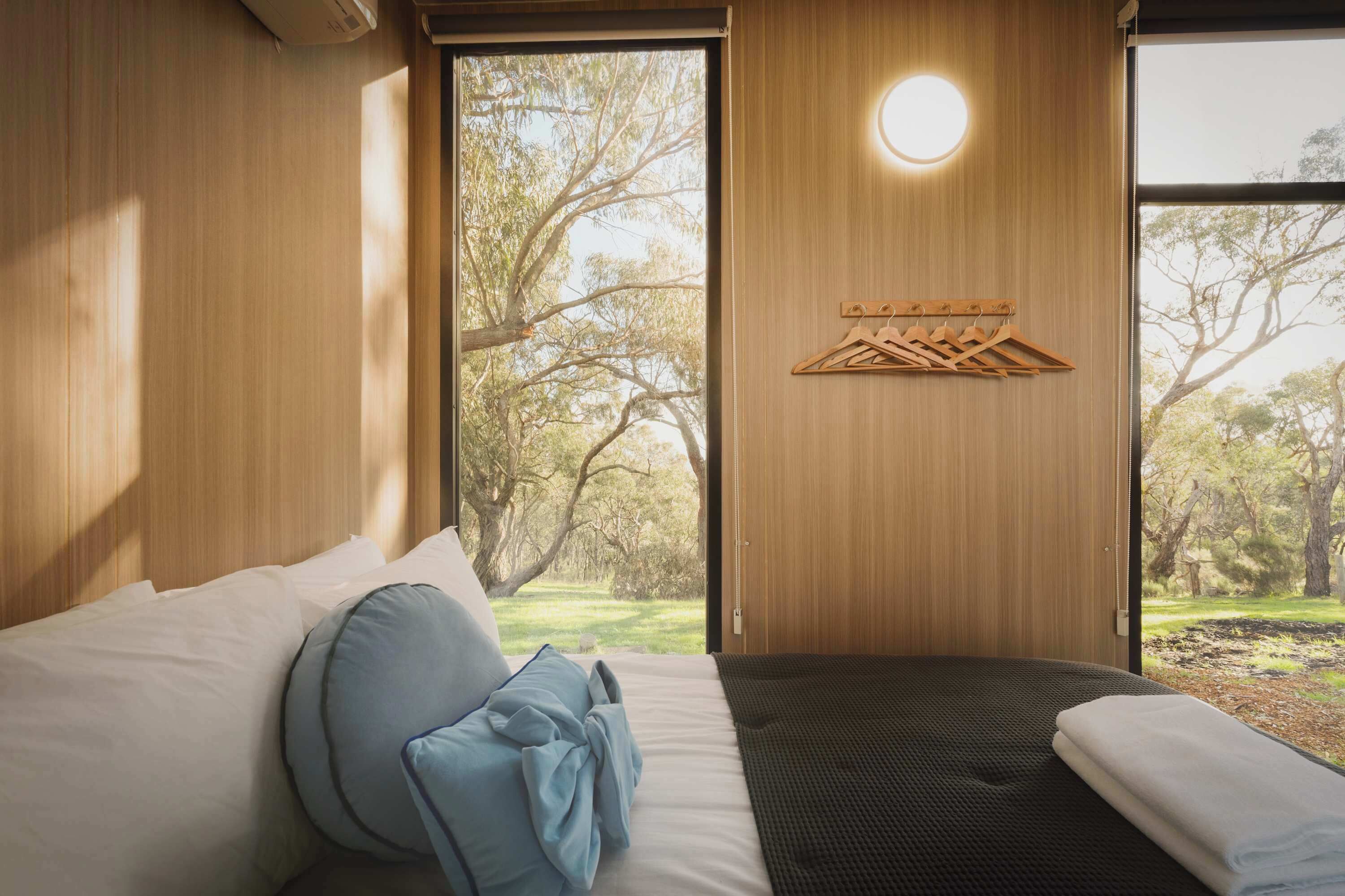 Een slaapkamer met een bed, een houten kapstok en uitzicht op bomen door een raam.