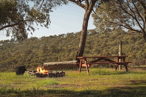 A picnic area with a fire, a wooden bench, and a picnic table in a grassy field with trees in the background.