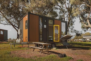 A small, modern cabin with a wooden exterior, a glass door, and a picnic table in a natural setting.