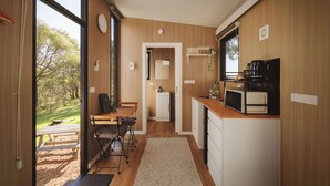 A compact kitchen with wooden cabinetry, a dining area with a table and chairs, and a view of the outdoors through large windows. - Tallarook Hilltop Tiny House 4 by Tiny Away (Tallarook)