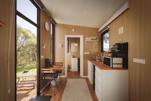 A compact kitchen with wooden cabinetry, a dining area with a table and chairs, and a view of the outdoors through large windows.