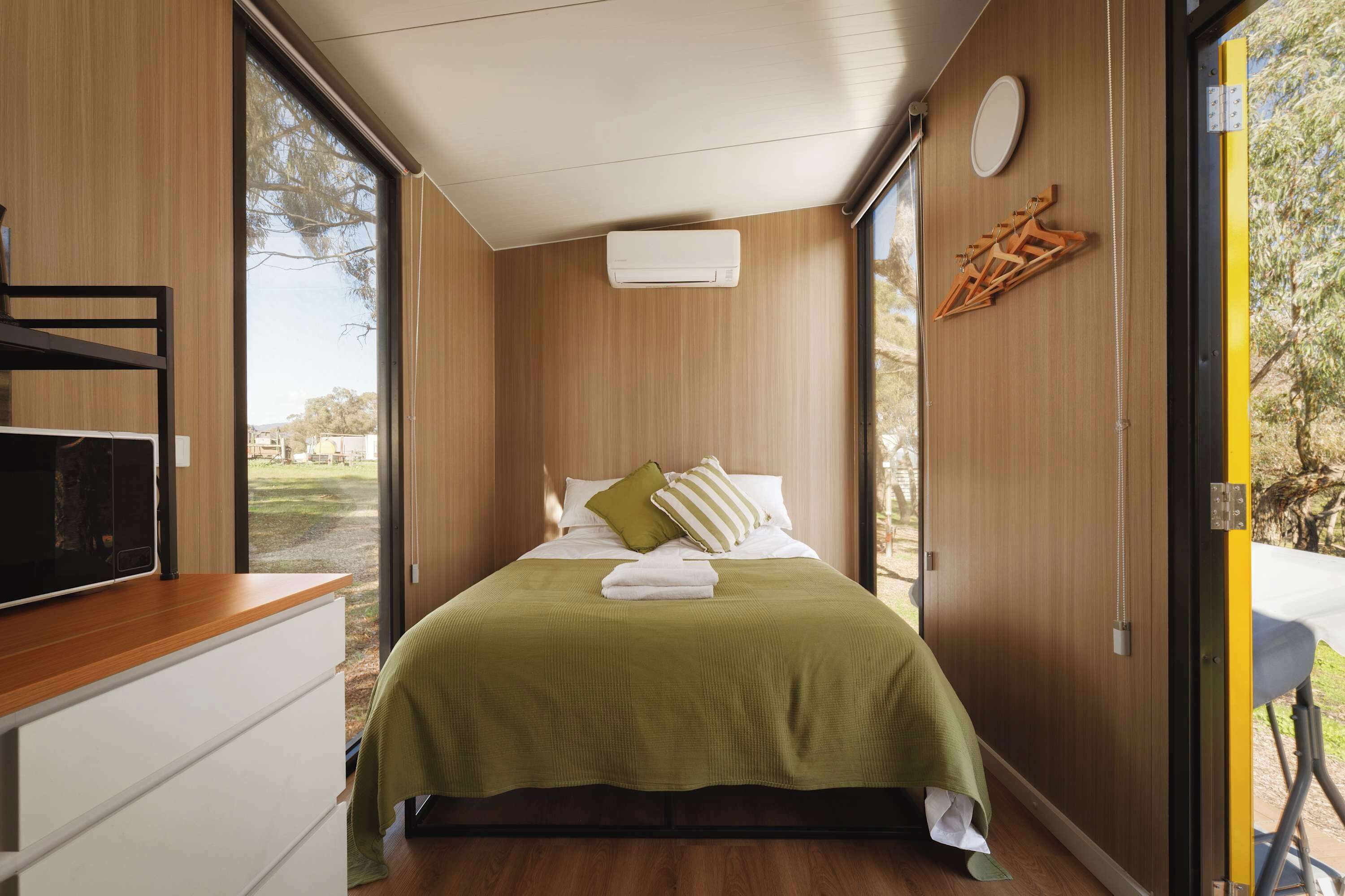 A bedroom with a bed, a bedside table, a wall-mounted air conditioner, and a view of the outdoors.