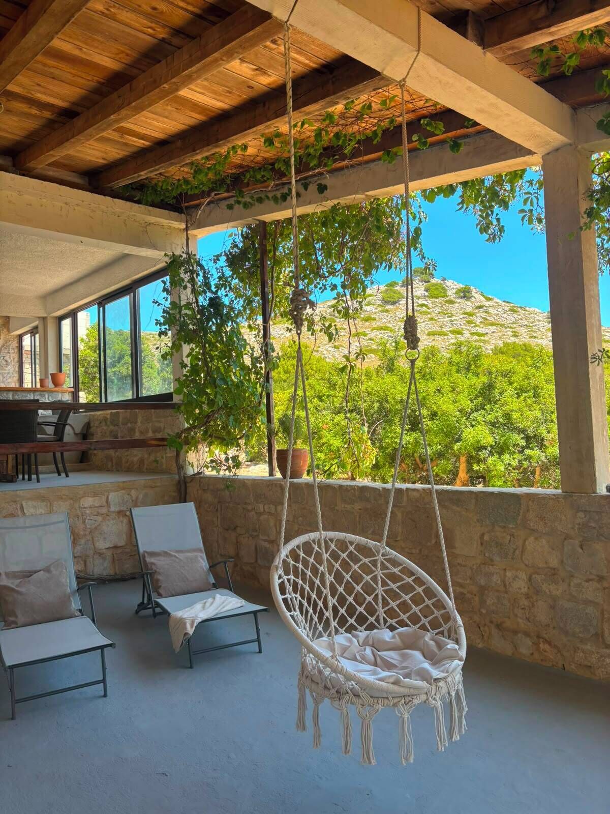 A covered outdoor seating area with a hanging chair, stone walls, and a view of greenery and hills.