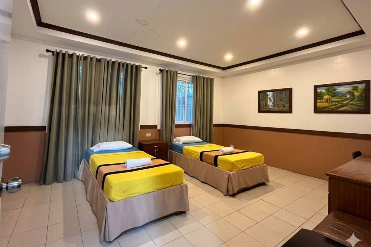 Ein Hotelzimmer mit zwei Einzelbetten, einem Schreibtisch, einem Stuhl, einem Fenster mit Vorhängen und einer Wand mit zwei gerahmten Bildern.