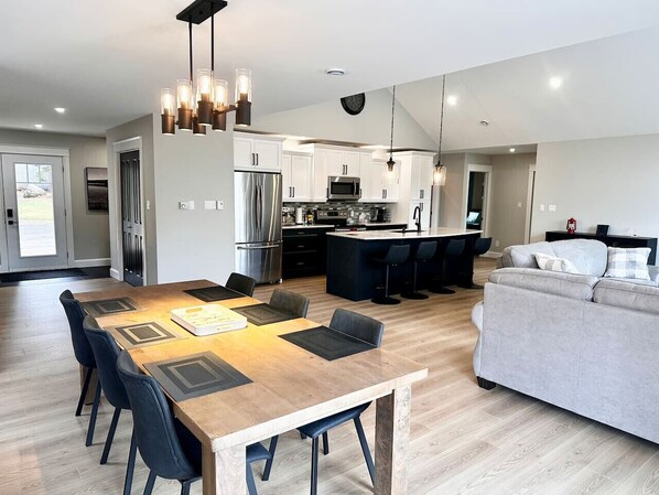 A modern kitchen with stainless steel appliances, a dining area with a wooden table and chairs, and a grey sofa in a spacious living area.