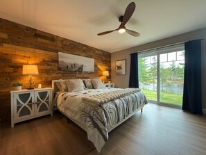 A bedroom with a bed, a nightstand, a ceiling fan, and a large window with a view of greenery.