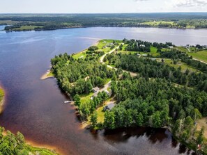 An aerial view of a lakeside property with a dock, surrounded by dense forest and a clear sky. - Waterfront | Kayaks | Screened Patio | Bonfire (Upper Rexton)