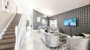 A modern living room with a staircase, a flat-screen TV, and a grey acoustic panel wall. - Disney Area 5BR With Pool Spacious and Prime Location (Kissimmee)