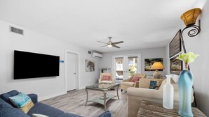 A living room with a sofa, a coffee table, a TV mounted on the wall, a ceiling fan, and a window with a view of the outdoors. - Ocean Breeze Escape South Padre Island (South Padre Island)
