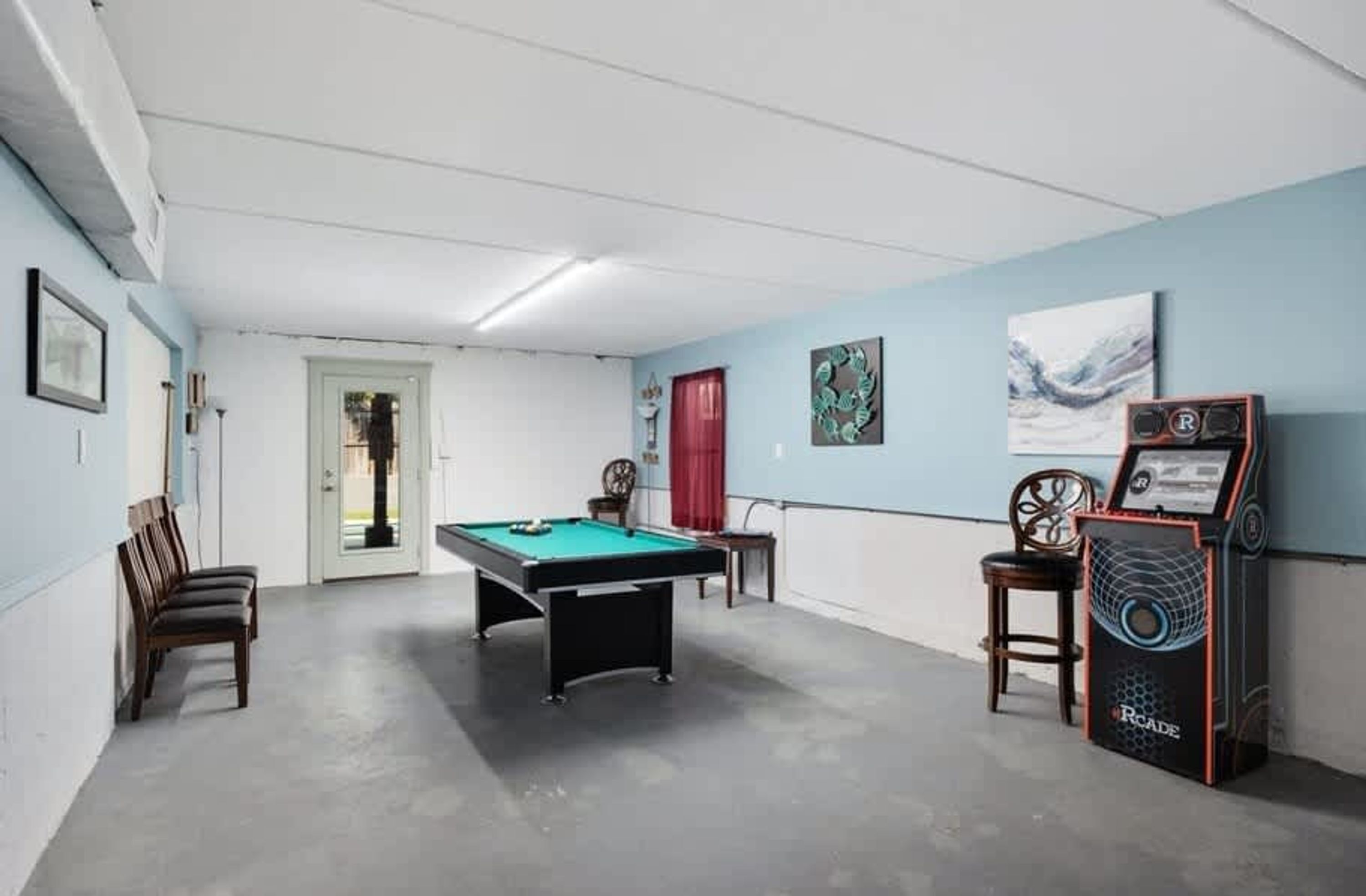 A room with a pool table, chairs, a door, and a video game machine.