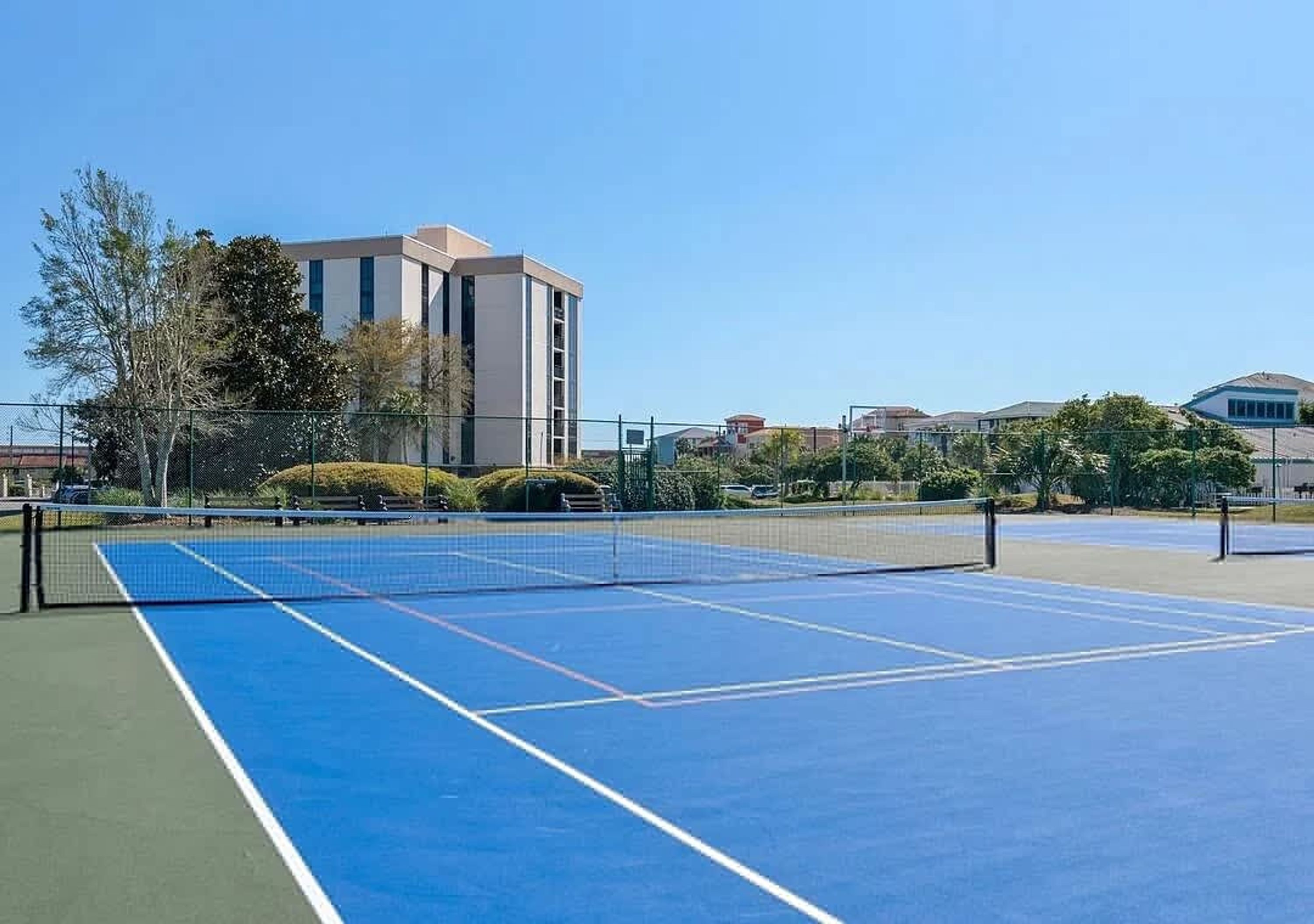 An outdoor tennis court with a net, surrounded by greenery and buildings.