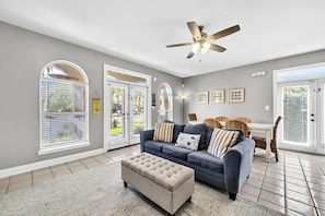 Un salon avec un canapé gris, un pouf beige, un ventilateur de plafond et un coin repas avec des chaises en osier.