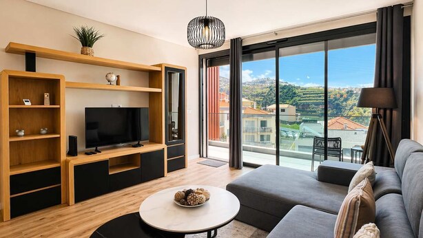 En moderne stue med fjernsyn, træhylder, sofa og balkon med udsigt over byen.
