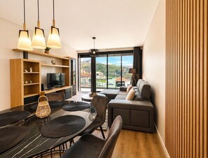 Ein modernes Wohnzimmer mit Essbereich, einer Sofagarnitur, einem Fernseher und einem großen Fenster mit Blick auf die Landschaft.