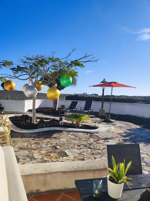 Una terraza en la azotea con un árbol, plantas en macetas, mobiliario exterior y un paraguas rojo.