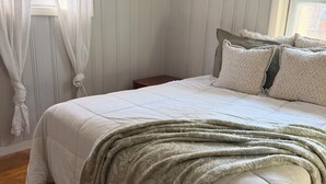 A bedroom with a bed, wooden flooring, a window with curtains, and a small bedside table.