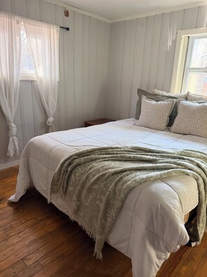 Ein Schlafzimmer mit einem Bett, Holzboden, einem Fenster mit Vorhängen und einem kleinen Nachttisch.