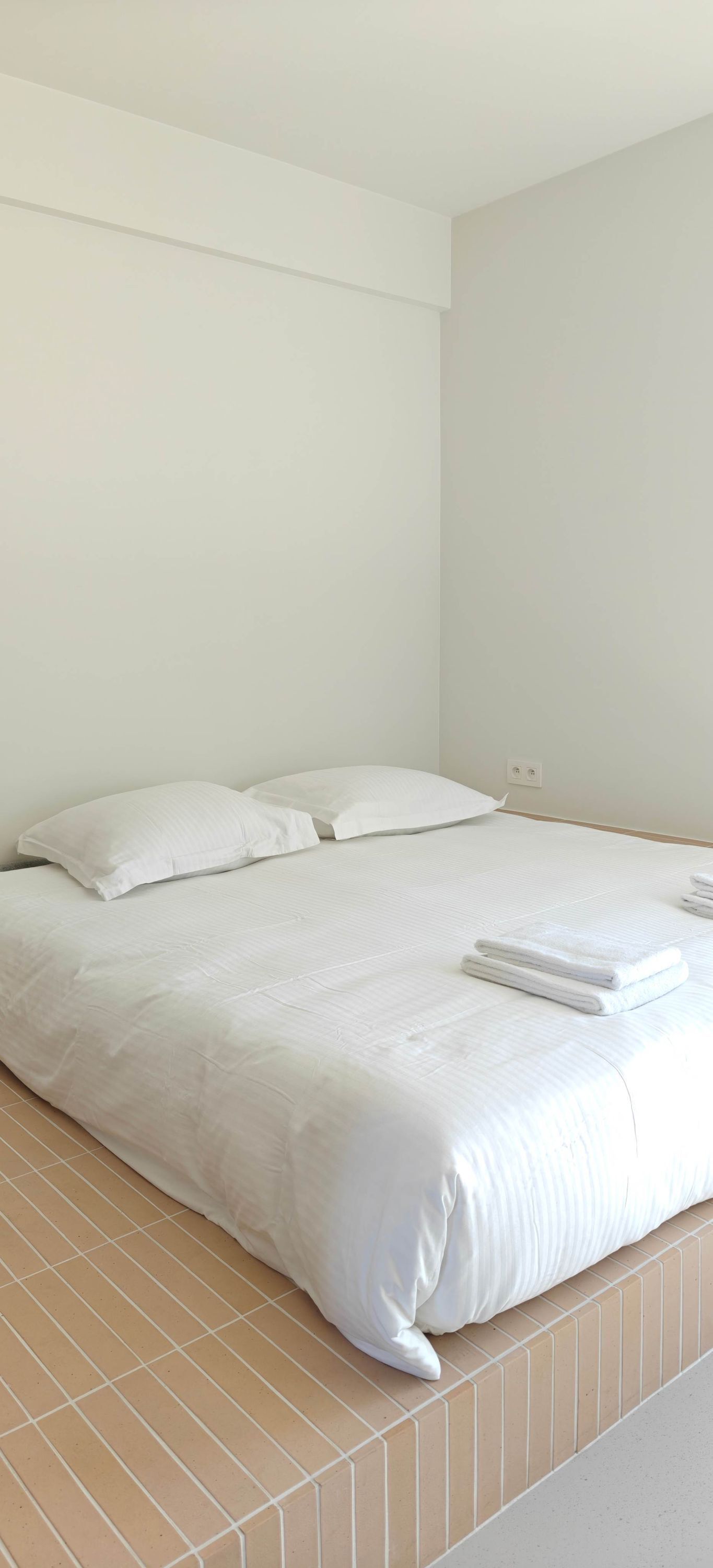 Ein ordentlich bezogenes Bett mit weißer Bettwäsche und Kissen, ein gefliester Boden und eine schlichte Wand.